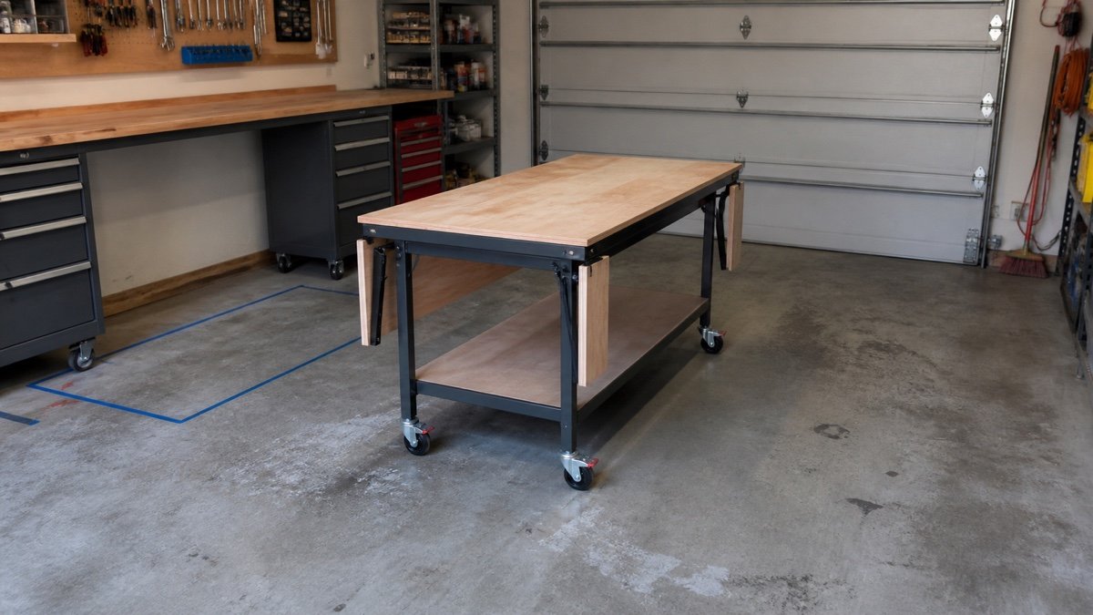 Mobile assembly table with locked casters beside its taped parking footprint in a garage workshop